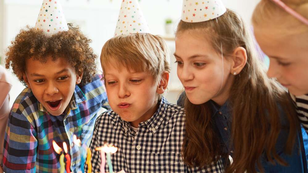 Kinder mit mit Geburtstagskappen pusten die Kerzen auf einem Kuchen aus.