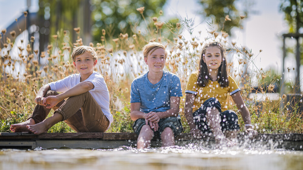 Drei Kinder sitzen am Rand des Becken vom Wasserspielplatz und plantschen mit den F&uuml;&szlig;en.