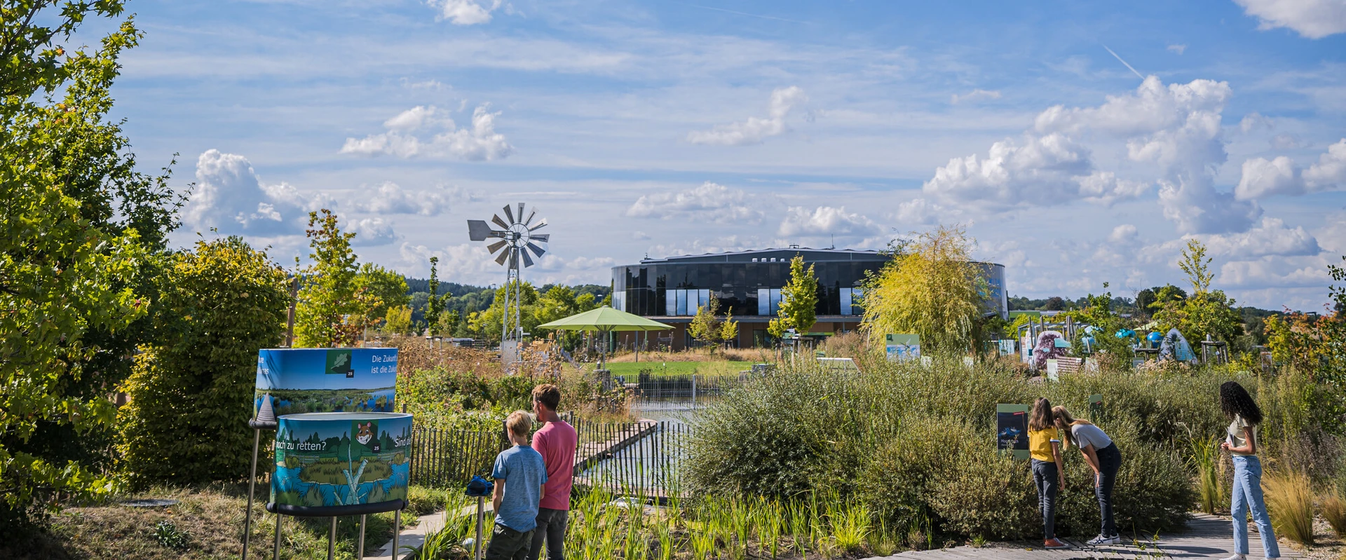 Blick vom angelegten Moor &uuml;ber den Themenpark auf das Geb&auml;ude der KLIMA ARENA in der Ferne. Besuchende auf Holzstegen &uuml;ber dem Moor.